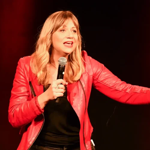 Comedian Mirja Regensburg in roter Lederjacke am Mikrofon vor dunkelrotem Bühnenhintergrund beim Cologne Comedy Festival.