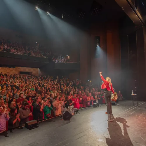 Komikerin Mirja Regensburg im roten Blazer winkt vom Bühnenrand ins stehend applaudierende, ausverkaufte Theater.