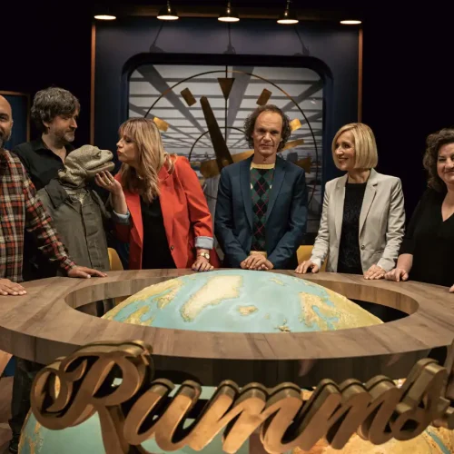 Komikerin Mirja Regensburg im roten Blazer mit dem Ensemble der Satire-Show „RUMMS!“ rund um den beleuchteten Globus-Tisch.