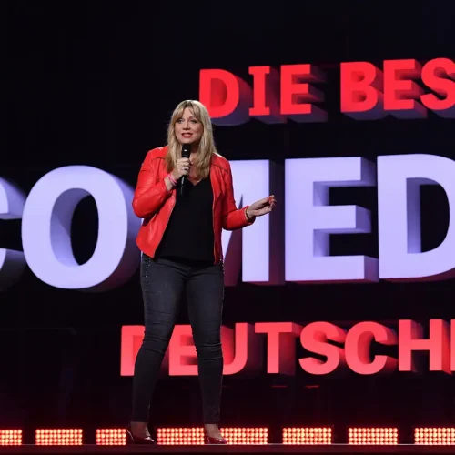 Comedian Mirja Regensburg in roter Lederjacke am Mikrofon vor dem riesigen Leuchtschriftzug „Die besten Comedians Deutschlands“.