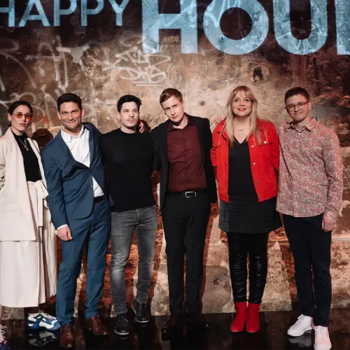 Komikerin Mirja Regensburg in roter Jacke mit der Gästerunde vor der leuchtenden „Happy Hour“-Wand der 3sat-Show.
