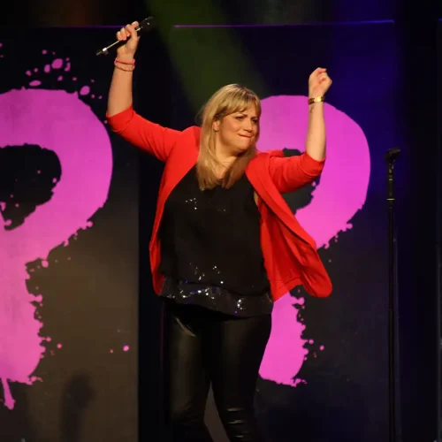Komikerin Mirja Regensburg tanzt gut gelaunt auf der Bühne vor einem pinken HAPPY-Hintergrund.