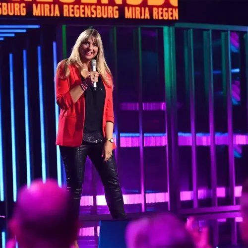 Komikerin Mirja Regensburg lacht mit Mikrofon auf der Bühne der Quatsch Comedy Show vor farbenfrohem Hintergrund.
