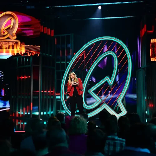Comedian Mirja Regensburg auf der großen Bühne der Quatsch Comedy Show vor dem riesigen leuchtenden türkisfarbenen Q-Logo.