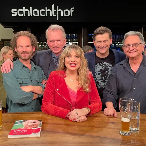 Komikerin Mirja Regensburg sitzt lächelnd am Holztisch im Münchener Schlachthof mit Michael Altinger und weiteren Kabarettisten.