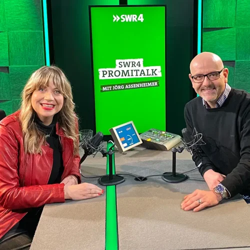 Komikerin Mirja Regensburg und Jörg Assenheimer lächeln gemeinsam am Mikrofon im SWR4-Podcaststudio.