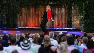 Komikerin Mirja Regensburg im roten Cardigan und roten Stiefeletten posiert vor dem Publikum auf der begrünten Open-Air-Bühne des ZDF Comedy Sommer.