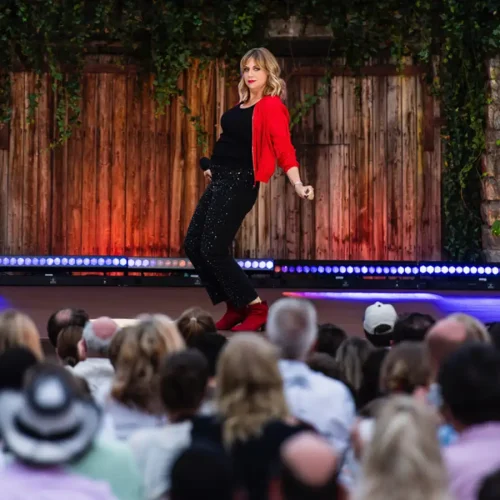 Komikerin Mirja Regensburg im roten Cardigan und roten Stiefeletten posiert vor dem Publikum auf der begrünten Open-Air-Bühne des ZDF Comedy Sommer.