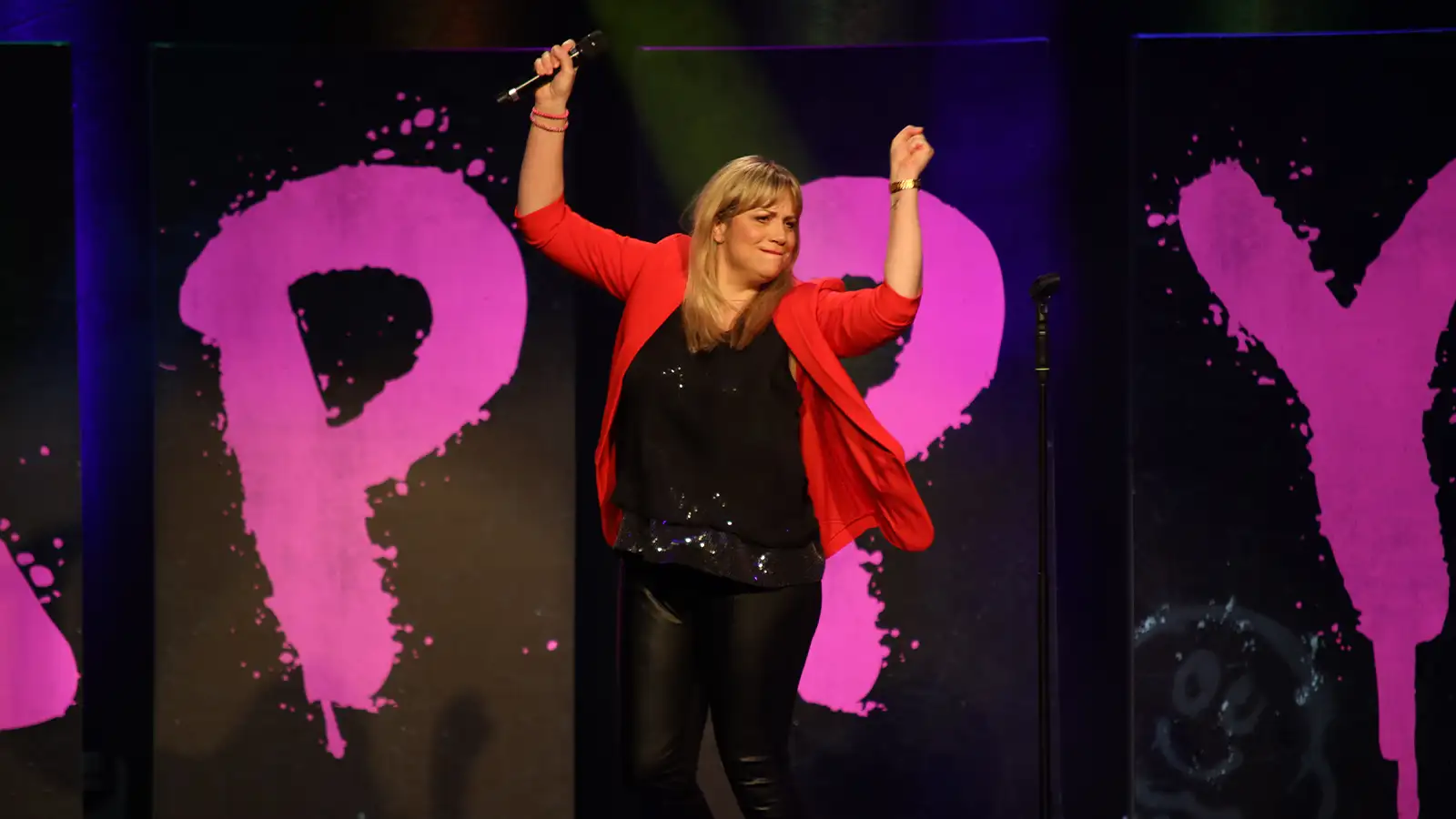 Mirja Regensburg tanzt gut gelaunt auf der Bühne vor einem pinken HAPPY-Hintergrund