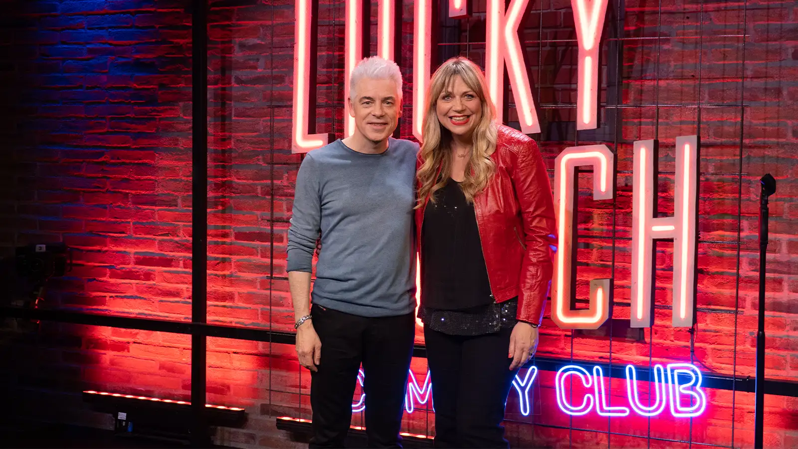 Mirja Regensburg im roten Sakko steht lächelnd neben Michael Mittermeier im Lucky Punch Comedy Club vor einer Backsteinwand mit Neon-Schriftzug