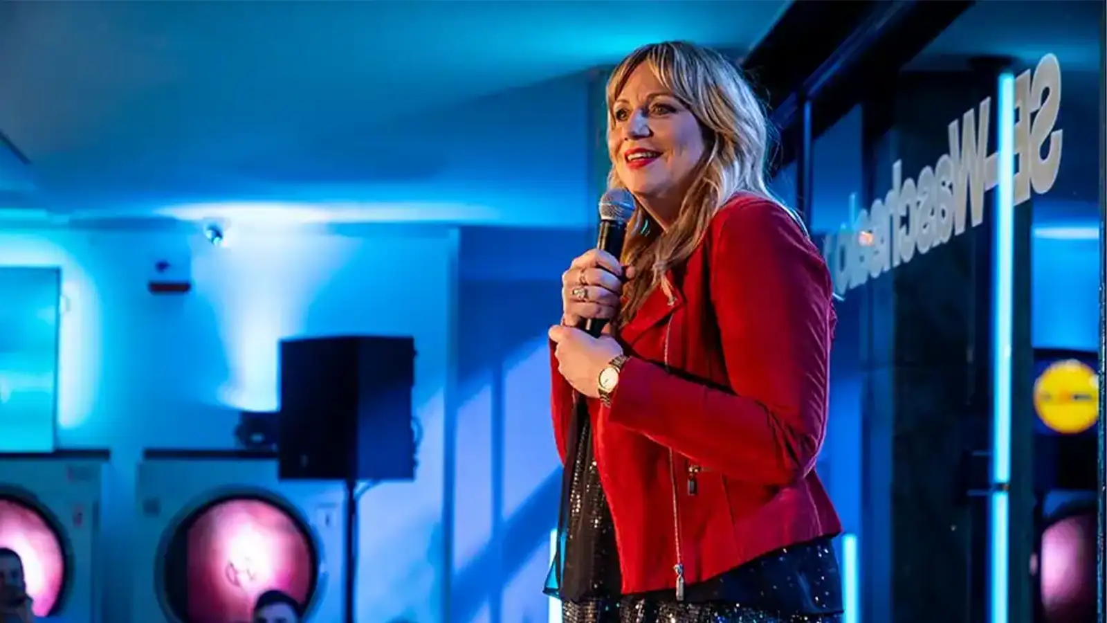 Mirja Regensburg moderiert mit Mikrofon im blau beleuchteten Eco-Express Waschsalon in Köln bei NightWash