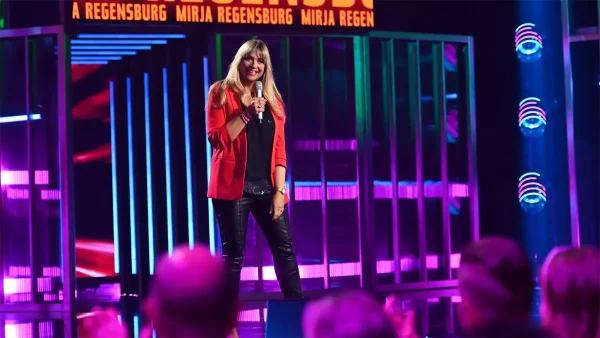 Mirja Regensburg lacht mit Mikrofon auf der Bühne der Quatsch Comedy Show vor einem farbenfrohen Hintergrund