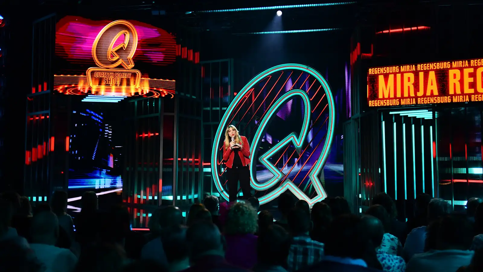 Mirja Regensburg steht auf der großen Bühne der Quatsch Comedy Show vor einem riesigen leuchtenden türkisfarbenen Q-Logo