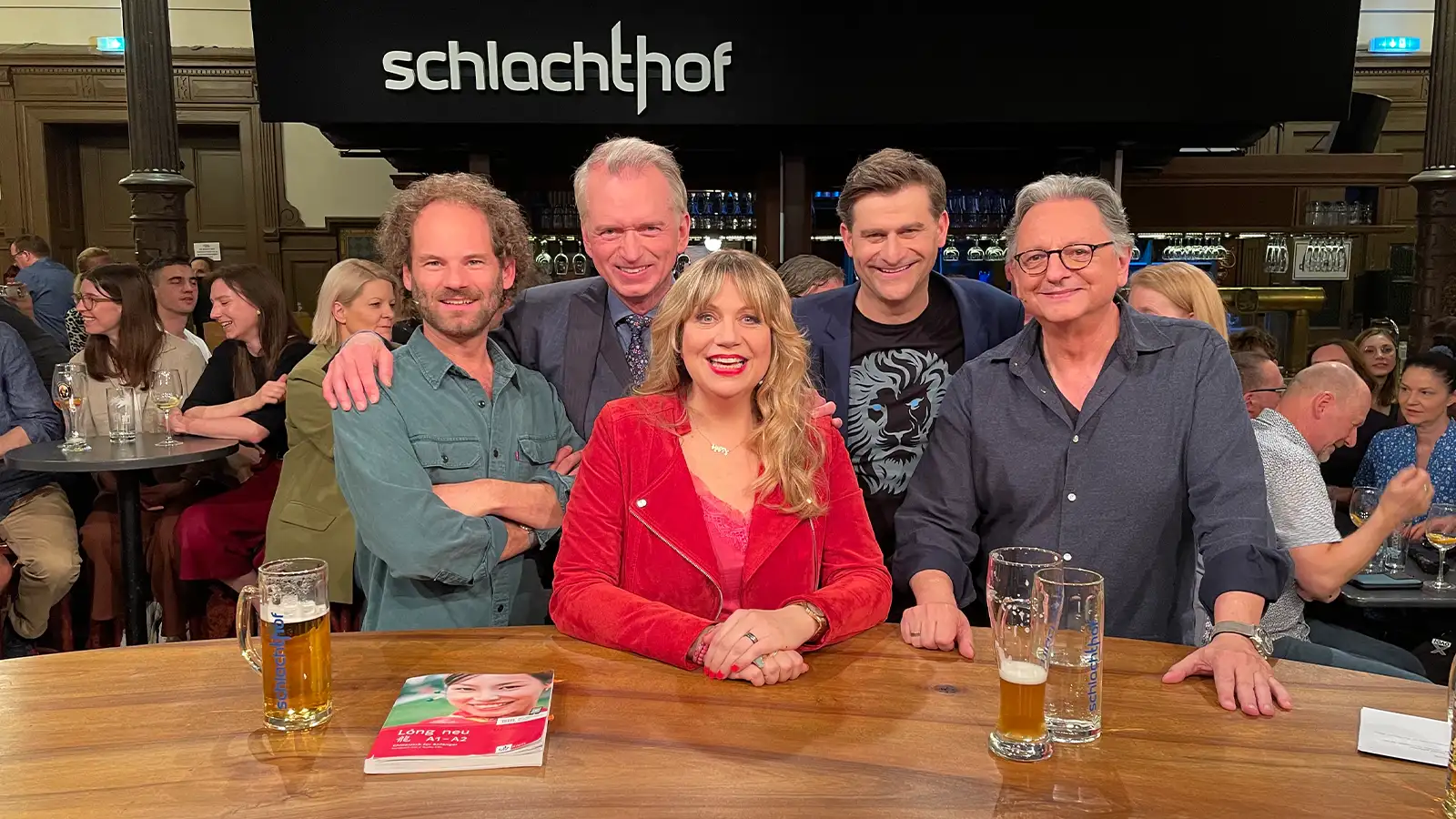 Mirja Regensburg sitzt lächelnd am hölzernen Talktisch im Münchener Schlachthof gemeinsam mit Michael Altinger und weiteren Kabarettisten