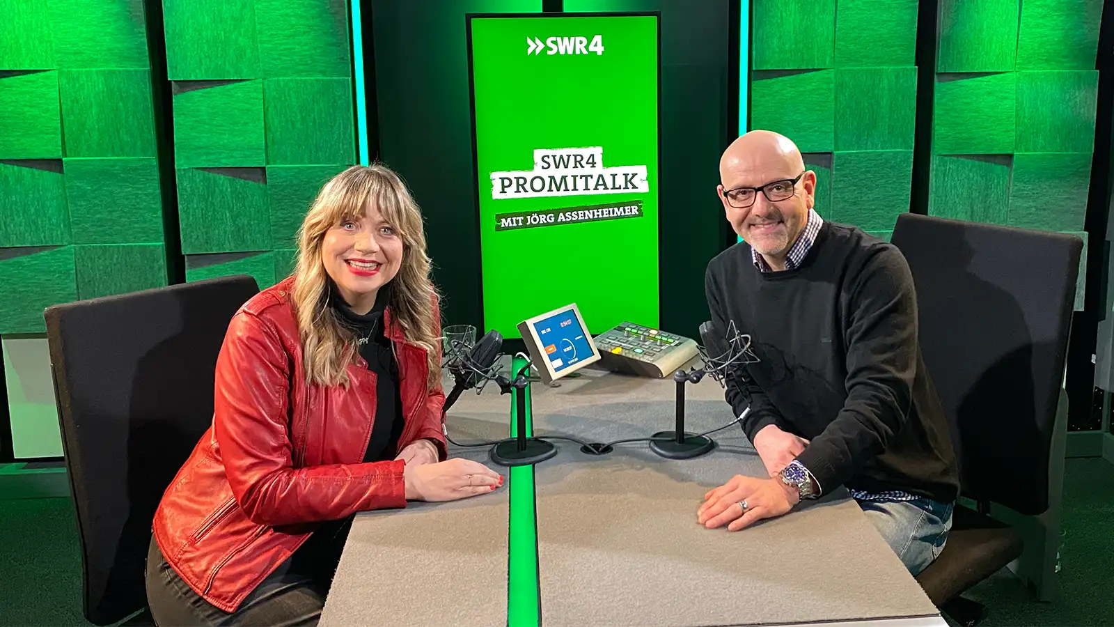 Mirja Regensburg und Jörg Assenheimer lächeln gemeinsam am Mikrofon im SWR4 Podcast-Studio